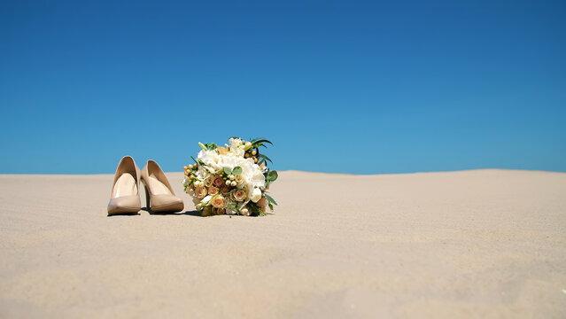 summer, desert, wedding female beige heels stand on the sand, next to the wedding bouquet. Day, against the blue sky. High quality photo