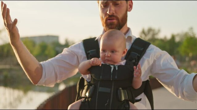 Tired young father with a little newborn baby in a slings carrier standing near lake at sunlight. Use phone talking to child on street. Ergo backpack. Parents. Gadget. Smartphone. Cell. Slow motion