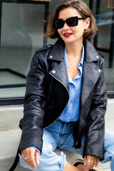 Close up portrait of pretty woman model wearing a black leather python jacket rock style, sunglasses, jeans sitting by street cafe.