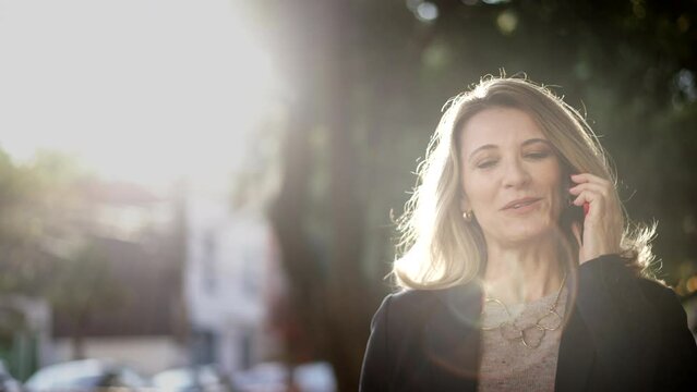 Happy successful middle aged female entrepreneur walking in street talking on phone. Businesswoman speaking on cellphone outdoors in sunlight