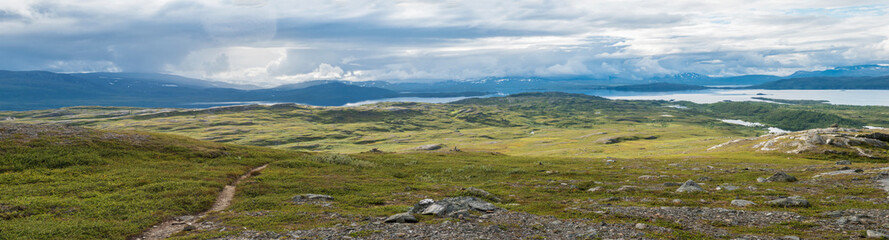 Naklejka na ściany i meble Panoramic landscape with beautiful Virihaure lake, snow capped mountain, birch trees and footpath of padjelantaleden hiking trail. Sweden, Lapland wild nature. Summer cloudy day