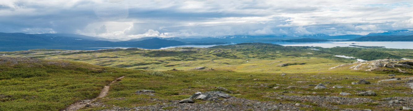 Panoramic landscape with beautiful Virihaure lake, snow capped mountain, birch trees and footpath of padjelantaleden hiking trail. Sweden, Lapland wild nature. Summer cloudy day