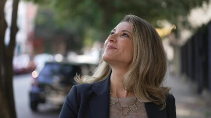 Carefree mature woman walking outdoors in city street in bright sunlight shining. 50s lady walking forward to camera - Powered by Adobe