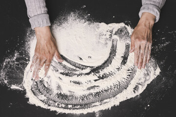 Sifted white wheat flour on a black table and two female hands, top view. cooking at home