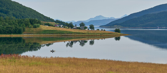 Morning landscape in Lofoten Island with symmetry reflection in the water of a lake. Natural mirror. Small fishing boat sailing on a lake. Mountain range at the background. Summer vacation in Lofoten.