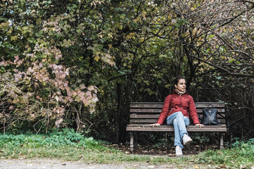 Portrait of woman sitting on bench in autumn park. Girl in red coat under tree branches