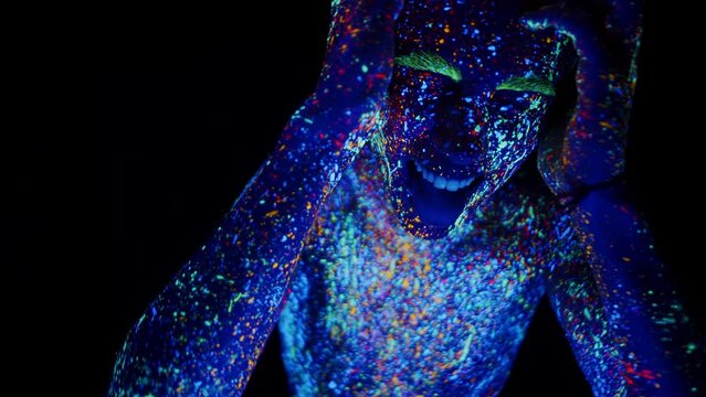 Young man in glowing neon bodyart holding his head in pain, panting, screaming in darkness. Concept of bad trip, drug abuse. Studio shot on black background