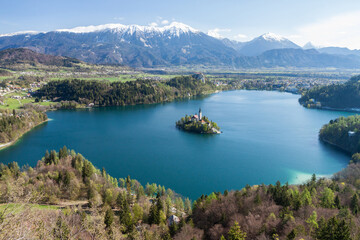 Fototapeta na wymiar Scenic, high elevation view of famous Lake Bled in Slovenia, with snowy Julian Alps mountain range in the background, spring growth on the trees and an island with a historical church.