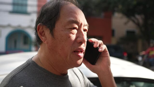One middle aged Asian man speaking on phone in street. Happy person listening on smartphone in sunlight