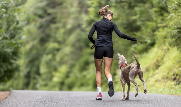 Picture from behind of a girl running in the nature with her dog jumping on her side