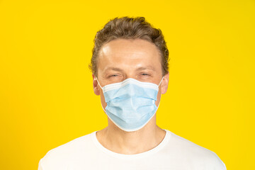 Happy, excited young man wearing a medical mask on his face. Sick young man in face mask wearing white t-shirt isolated on yellow background. A smiling caucasian man in protective face mask