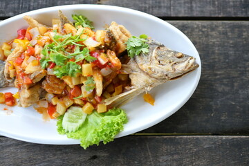 Deep-Fried snapper with Three-Flavoured Sauce. Deep Fried Fish with Chili Sauce, Pineapple, onion, tomato, carrot (all diced) topped with shredded coriander served in a white plate on a wood table.
