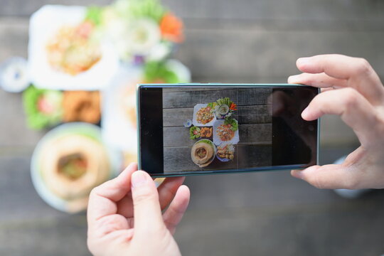 A woman holding a smartphone or mobile phone takes pictures of a set Thai dishes and shares it on social networks before eating. Food served on wooden table consists of vermicelli salad,steamed,etc. 
