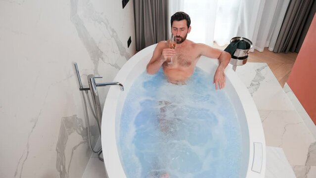 Rich happy sexy businessman enjoying jacuzzi spa in hotel after well done job drinking champagne. Portrait of handsome guy relaxing in in the bath. Attractive man having hydro massage indoor. 