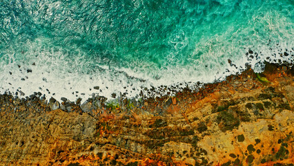 Drone view over azure ocean water and rocky shore. Top view over rolling ocean waves to the rocky shore. Aerial view of the beautiful ocean and rocky shore with rolling waves, on summer day.
