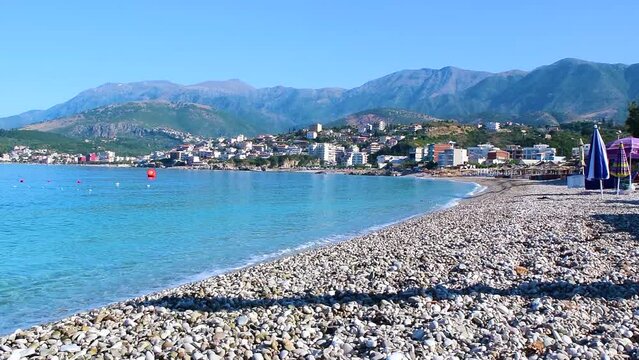 Beautiful pebble beach of Himare town at foot of mountains. Ionian sea. Albania. Summer sunny landscape. Concept of summer holidays and relaxation.