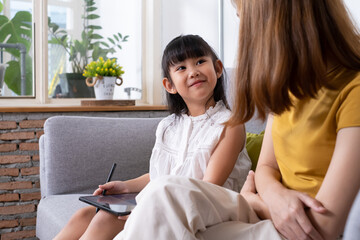 Young Asian girl using digital tablet for painting picture or study online lesson at home, with mother support and teaching the lesson inside of living room. Online education or home school concept.