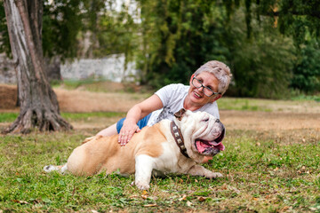 Smiling senior woman laying on grass with english bulldog outdoors. Woman hugging dog in park
