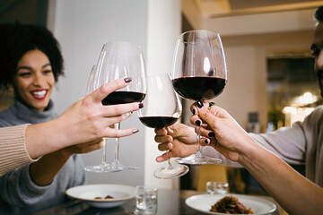 Happy multiracial best friends dining at restaurant and celebrating with a red wine toast together - crop shot details on the hands holding wineglasses - people, food and drink lifestyle concept