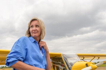 Fototapeta premium Travel in bad weather. A beautiful blonde woman in a blue shirt against the background of a whitish yellow plane in against the backdrop of storm clouds