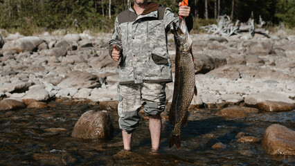 male fisherman holds a large fish pike caught on the shore of the lake against the background of stones