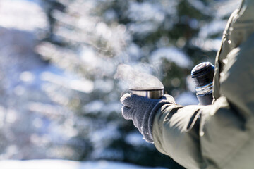 Winter picnic in snowy forest. Man drinking coffee from thermos. Traveler pouring steamy tea and holding metal cup with hot beverage. Lifestyle moment in nature. Close up of hands getting warm