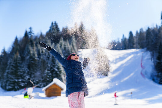 Happy child throwing snow on sunny winter day outdoor. Kid having fun at snowy ski resort, enjoying Christmas holidays in mountain. Girl turn snowflakes in magical glitter. Authentic lifestyle moment