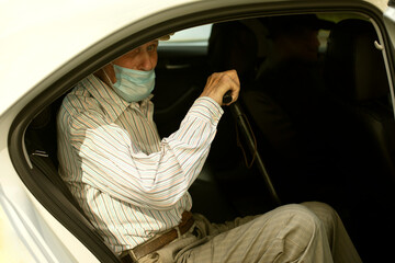 An old man in a taxi. A man in a medical mask is sitting in a car. Outside view of the pensioner sitting in the car.