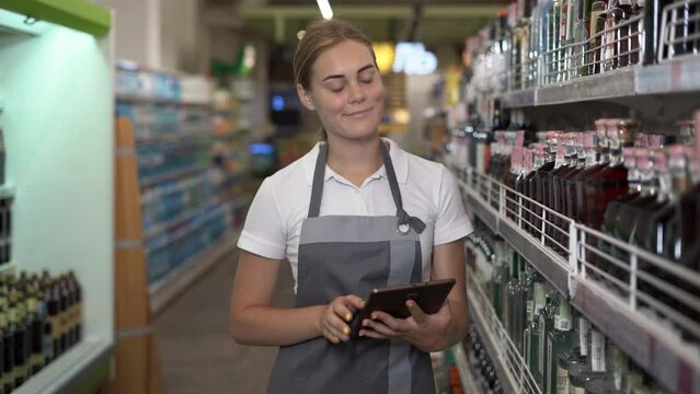 A successful worker in a store in a casual gray apron, walking between the shelves with alcohol in a supermarket and holding tablet in his hands. Concept of young woman trainee.