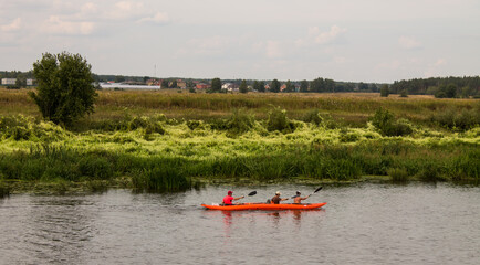 Three tourists are sailing on an orange kayak on the Klyazma river in the Moscow region among the banks with green grass