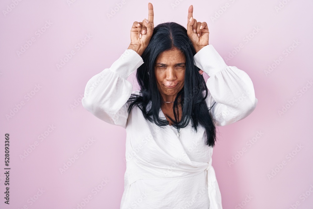 Poster mature hispanic woman standing over pink background doing funny gesture with finger over head as bul