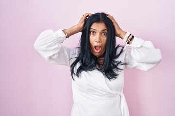 Mature hispanic woman standing over pink background crazy and scared with hands on head, afraid and surprised of shock with open mouth