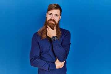 Redhead man with long beard wearing casual blue sweater over blue background looking confident at the camera smiling with crossed arms and hand raised on chin. thinking positive.