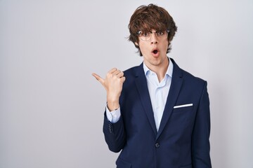 Hispanic business young man wearing glasses surprised pointing with hand finger to the side, open mouth amazed expression.