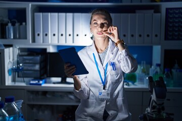 Beautiful blonde woman working at scientist laboratory late at night mouth and lips shut as zip...