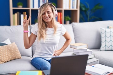 Young blonde woman studying using computer laptop at home smiling looking to the camera showing fingers doing victory sign. number two.