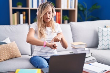 Young blonde woman studying using computer laptop at home in hurry pointing to watch time,...