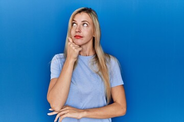 Beautiful blonde woman wearing casual t shirt over blue background with hand on chin thinking about question, pensive expression. smiling with thoughtful face. doubt concept.
