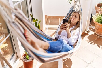 Young blonde woman using smartphone lying on hammock at terrace.