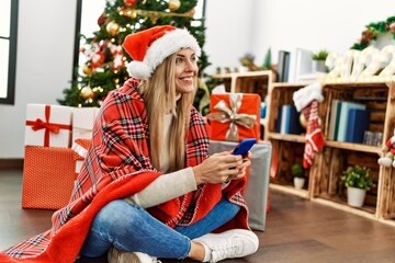 Young blonde woman using smartphone sitting by christmas tree at home