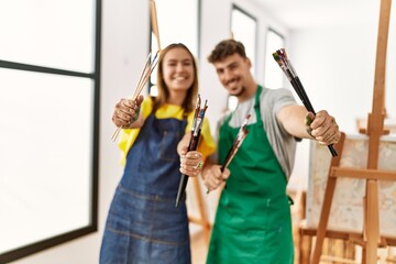 Young hispanic artist couple smiling happy holding paintbrushes at art studio.