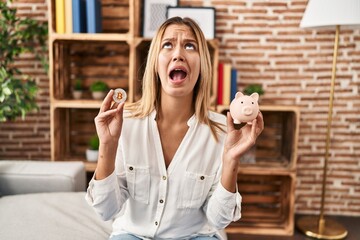 Young blonde woman holding piggy bank and bitcoin angry and mad screaming frustrated and furious,...