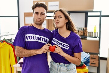 Young hispanic couple wearing volunteer t shirt holding heart clueless and confused expression. doubt concept.