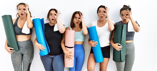 Obraz premium Group of women holding yoga mat standing over isolated background doing ok gesture shocked with surprised face, eye looking through fingers. unbelieving expression.