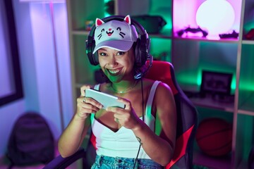 Young beautiful hispanic woman streamer playing video game by smartphone at gaming room