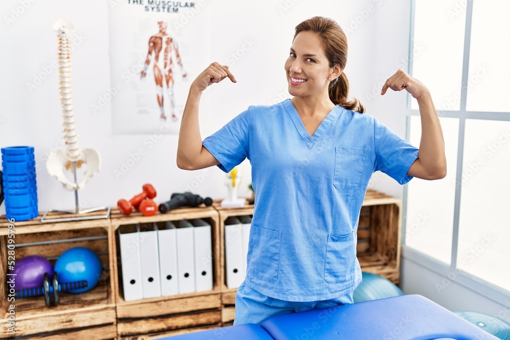 Wall mural Middle age hispanic physiotherapist woman working at pain recovery clinic looking confident with smile on face, pointing oneself with fingers proud and happy.