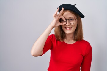 Young redhead woman standing wearing glasses and beret doing ok gesture with hand smiling, eye looking through fingers with happy face.
