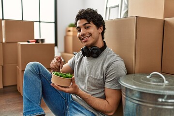 Obraz na płótnie Canvas Young hispanic man eating salad sitting on the floor at new home.