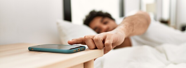 Young hispanic man turning off smartphone alarm lying on the bed at bedroom.