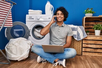 Young hispanic man using laptop and talking on the smartphone waiting for washing machine at laundry room
